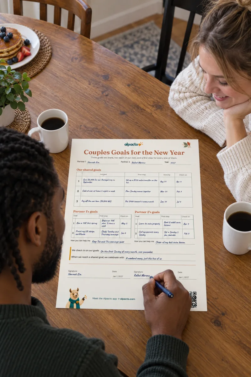 A couple at a wooden kitchen table with the filled-in couples goal setting worksheet, warm design; he holds a pen at the second signature line while she watches
